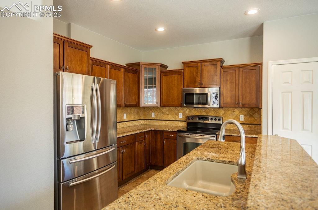 Kitchen with stainless steel appliances, glass fronted cabinets, light stone counters, wood finish cabinetry, and tasteful backsplash