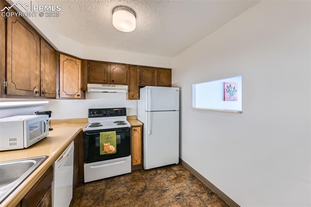 Kitchen featuring white appliances, under cabinet range hood, a textured ceiling, light countertops, and dark stone finish floors