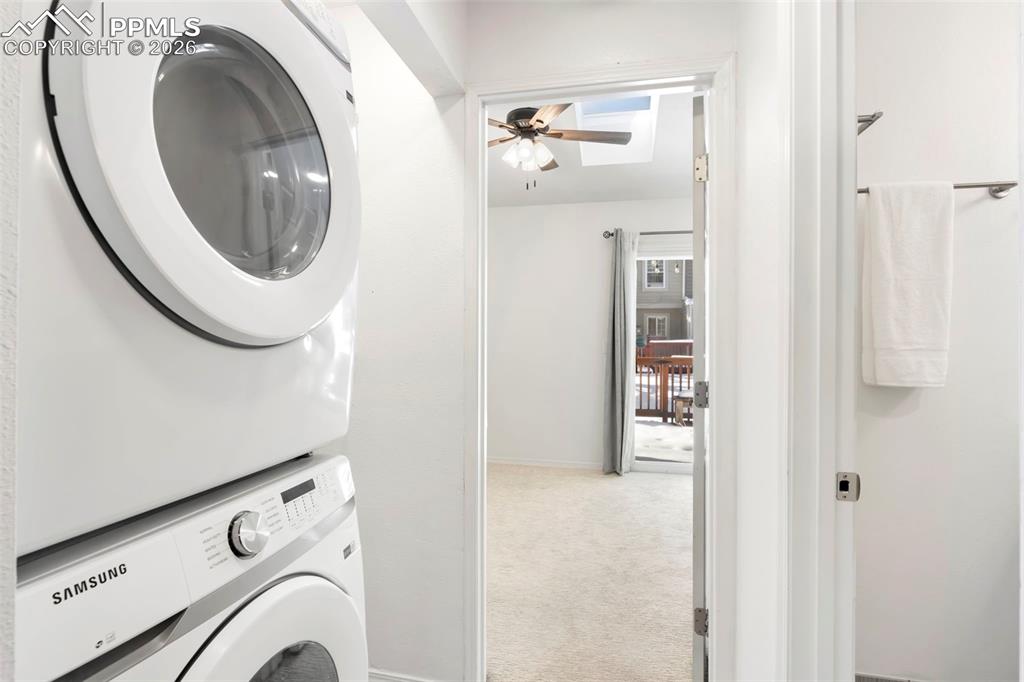 Laundry area featuring light colored carpet, stacked washing machine and dryer, and ceiling fan