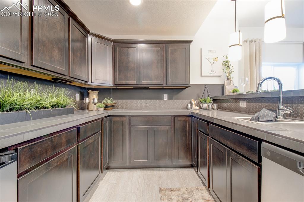 Kitchen featuring backsplash, tile countertops, dishwasher, dark brown cabinetry, and pendant lighting