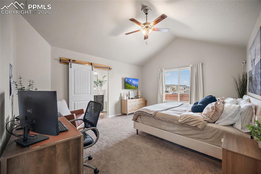 Carpeted bedroom featuring an office area, a barn door, ceiling fan, ensuite bath, and lofted ceiling