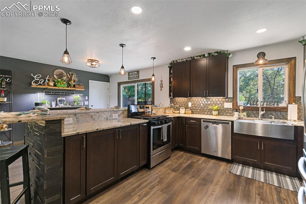 Kitchen with dark brown cabinets, a peninsula, appliances with stainless steel finishes, light stone counters, and dark wood-style floors