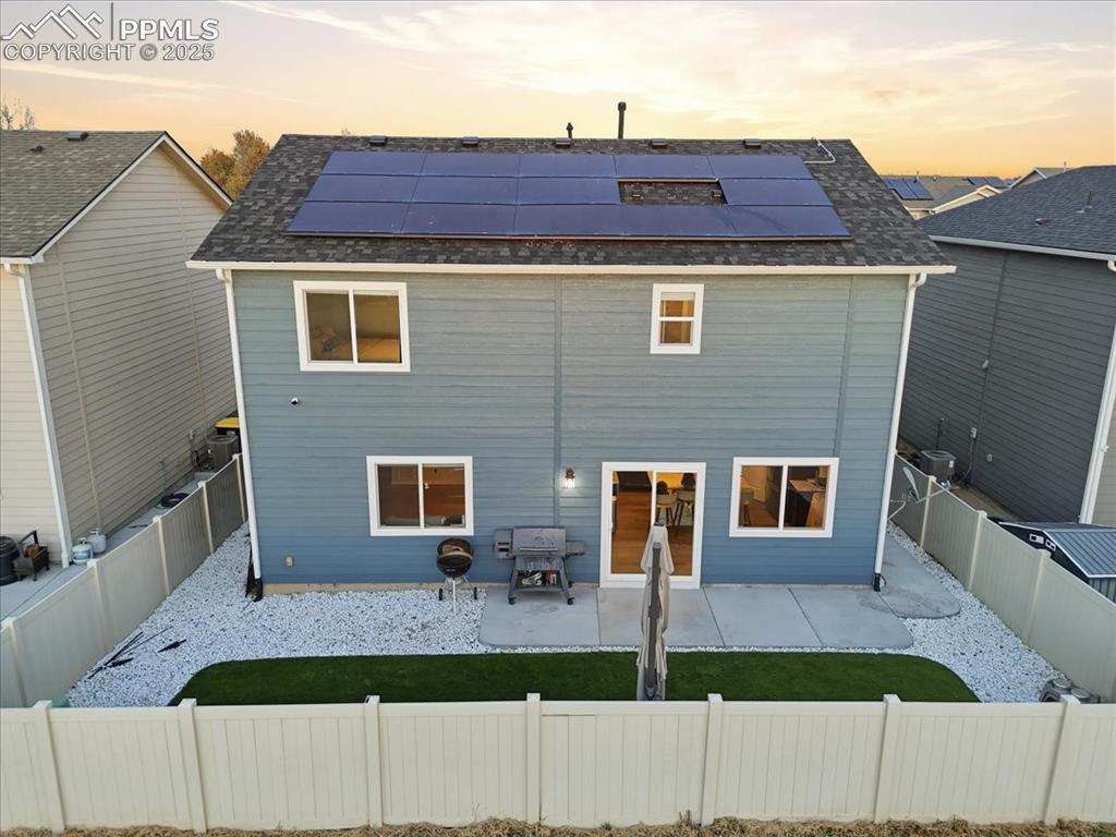 Back of property at dusk with a patio, roof mounted solar panels, a fenced backyard, and a shingled roof