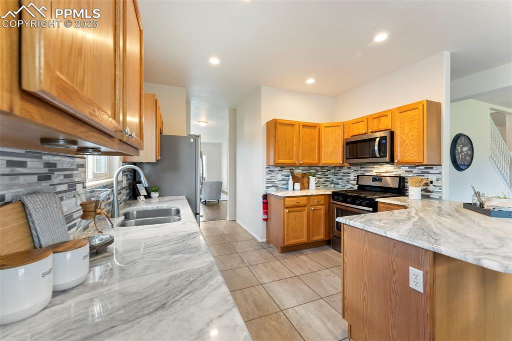 Kitchen featuring backsplash, brown cabinetry, light stone countertops, stainless steel appliances, and light tile patterned floors