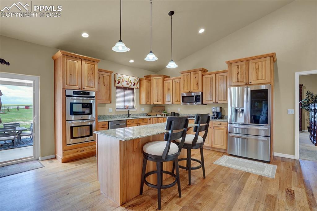 Kitchen with hanging light fixtures, appliances with stainless steel finishes, light wood-style floors, a kitchen island, and light stone countertops