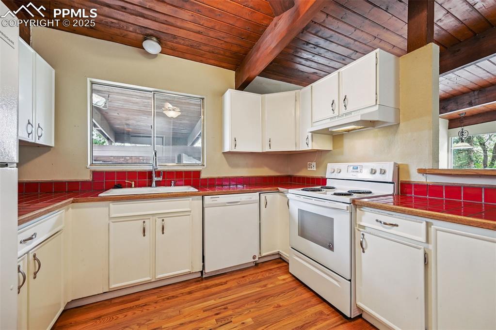 Kitchen w/wood ceiling and exposed beams, white appliances, wood floors, under cabinet range hood, and white cabinetry