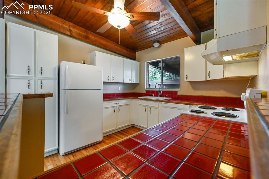 Kitchen featuring tile counters, a wood ceiling with exposed beams, white appliances, white cabinets, and under cabinet range hood