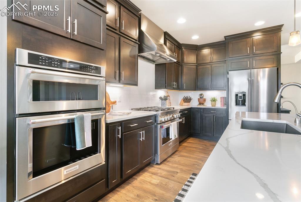 Kitchen featuring premium appliances, wall chimney exhaust hood, light wood finished floors, light stone countertops, and decorative backsplash