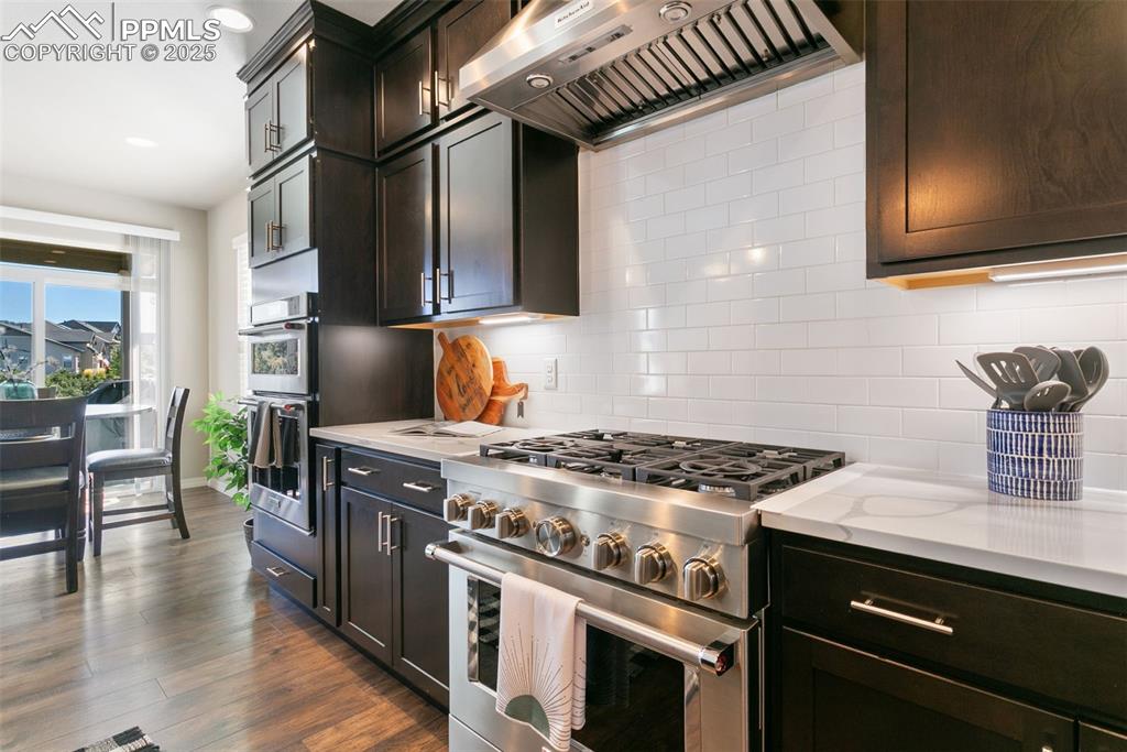 Kitchen with appliances with stainless steel finishes, wall chimney range hood, decorative backsplash, dark wood-style floors, and light stone counters