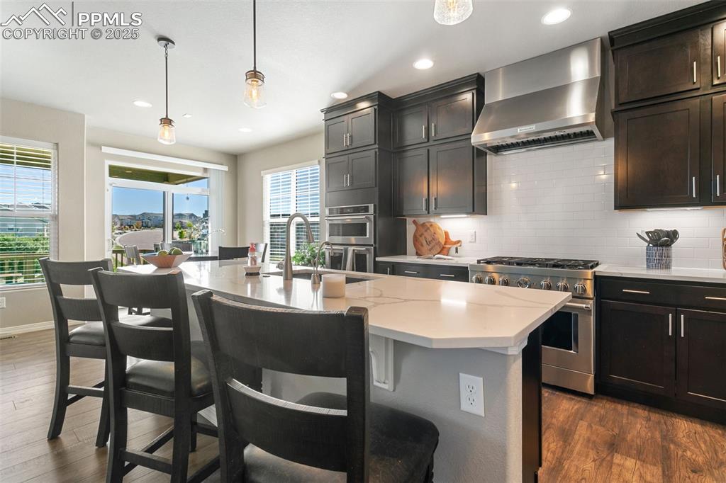 Kitchen with pendant lighting, decorative backsplash, wall chimney exhaust hood, appliances with stainless steel finishes, and dark wood-style flooring