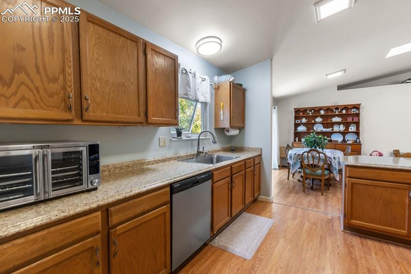 Kitchen with brown cabinets, light wood-style flooring, stainless steel dishwasher, light stone counters, and vaulted ceiling