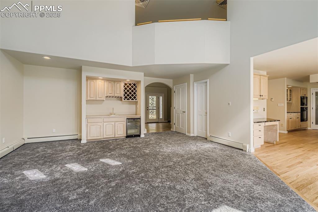 Unfurnished living room with arched walkways, baseboard heating, bar with sink, a high ceiling, and beverage cooler (main floor)