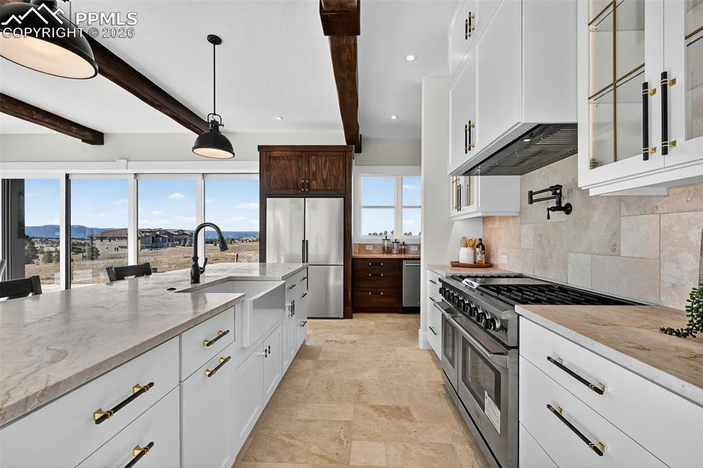 Two tone kitchen with stainless steel appliances, light stone countertops, beamed ceiling, glass insert cabinets, and pendant lighting