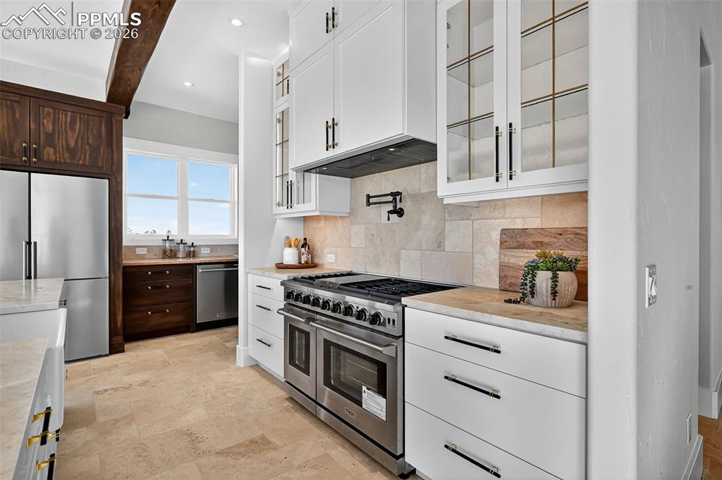 Kitchen featuring stainless steel appliances, glass fronted cabinets, light stone counters, beam ceiling, and tasteful backsplash