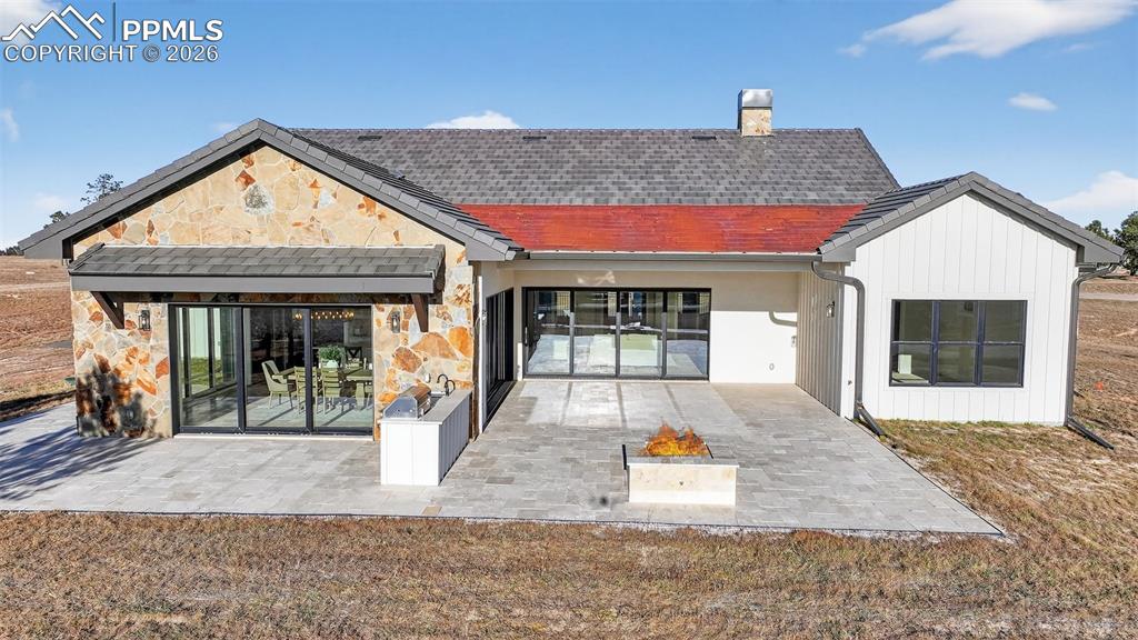 Back of house with stone siding, a chimney, a patio area, and roof with shingles