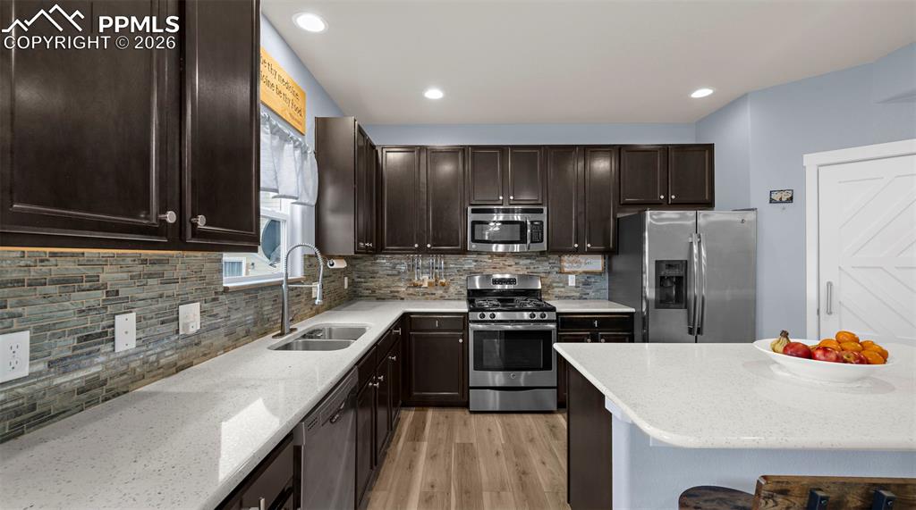 Kitchen with dark wood finish cabinets, stainless steel appliances, light stone counters, light wood-style floors, and backsplash