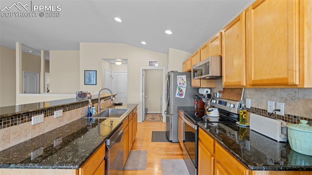 Kitchen featuring decorative backsplash, stainless steel appliances, light wood-type flooring, vaulted ceiling, and dark stone countertops