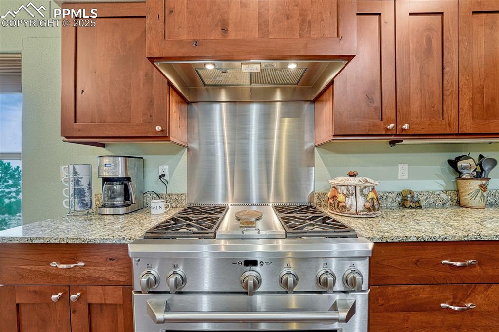 Kitchen featuring high end range, brown cabinetry, light stone countertops, and range hood