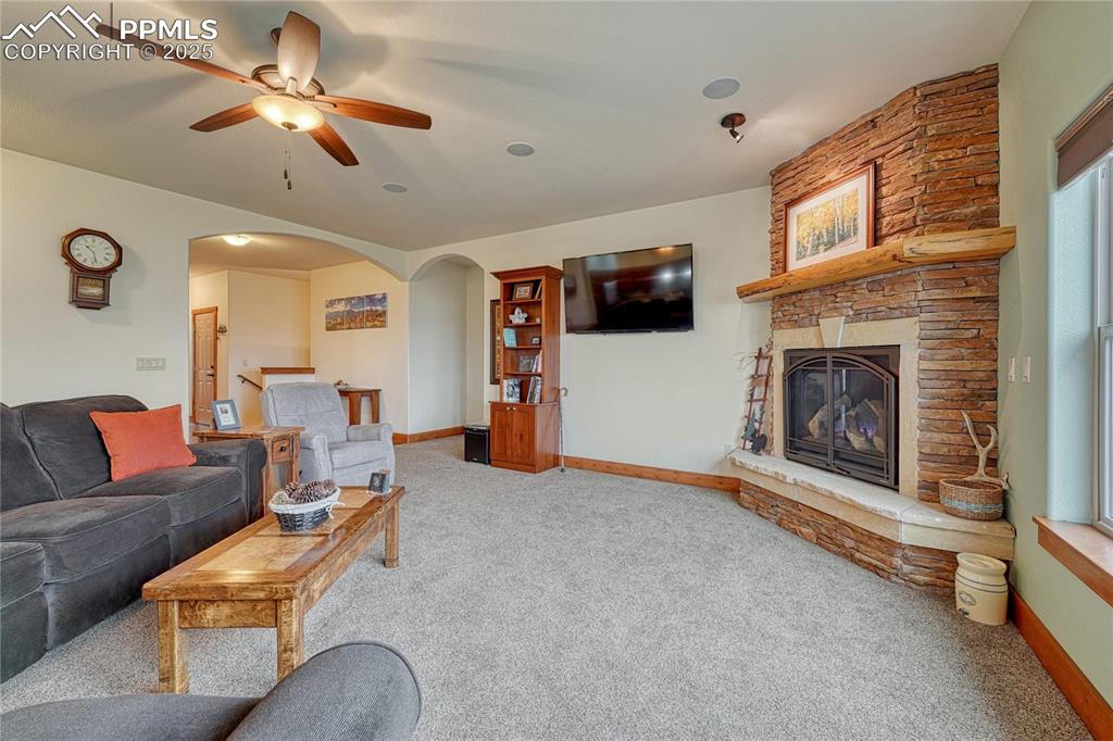 Living room featuring carpet floors, arched walkways, a ceiling fan, a stone fireplace, and baseboards