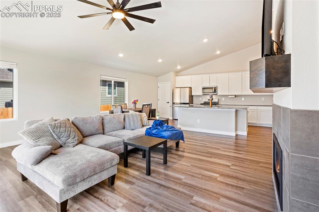 Living room with recessed lighting, lofted ceiling, light wood finished floors, a ceiling fan, and a warm lit fireplace