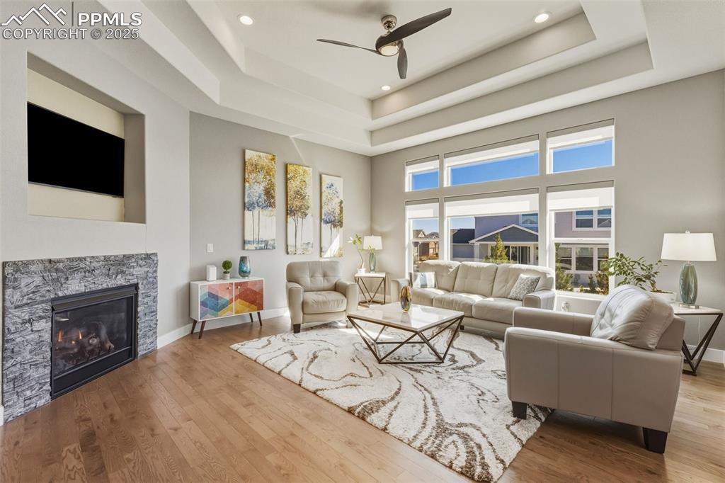 Living area featuring a ceiling fan, a raised ceiling, a glass covered fireplace, wood finished floors, and recessed lighting
