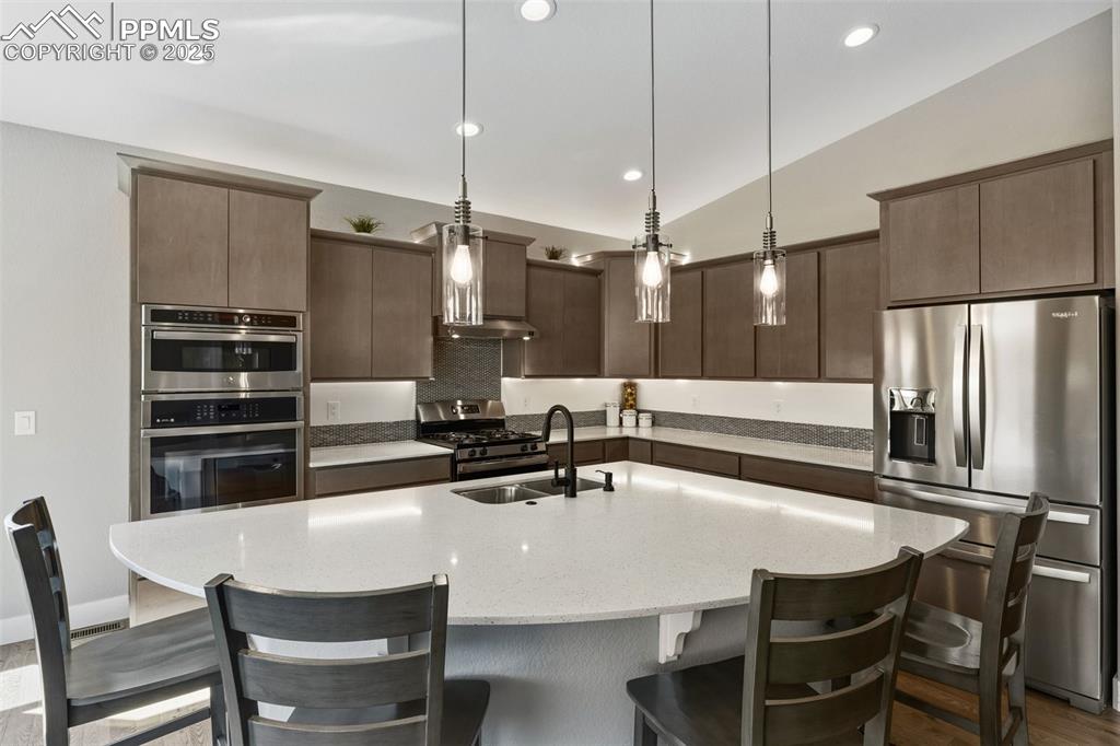 Kitchen featuring stainless steel appliances, decorative light fixtures, a center island with sink, recessed lighting, and light stone countertops