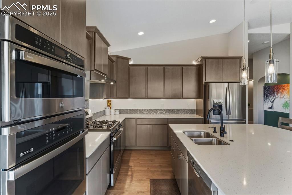 Kitchen with stainless steel appliances, vaulted ceiling, light wood-style floors, pendant lighting, and light stone counters