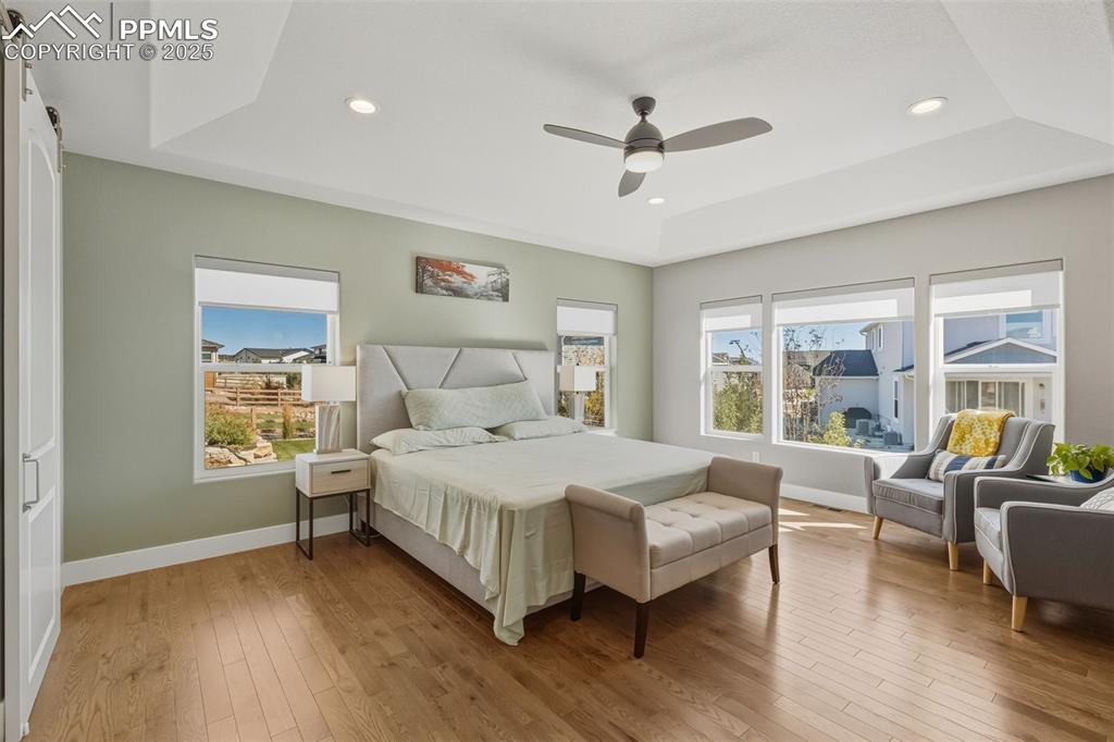Bedroom with a tray ceiling, light wood-style floors, a barn door, ceiling fan, and recessed lighting