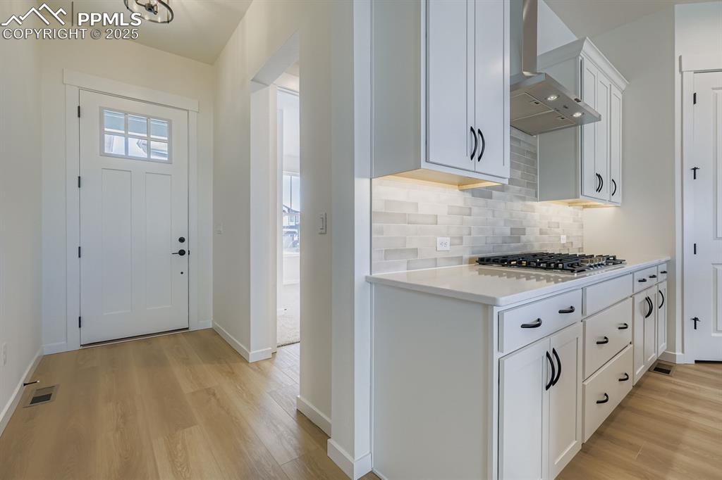 Kitchen with white cabinetry, light wood-type flooring, wall chimney exhaust hood, tasteful backsplash, and light stone countertops