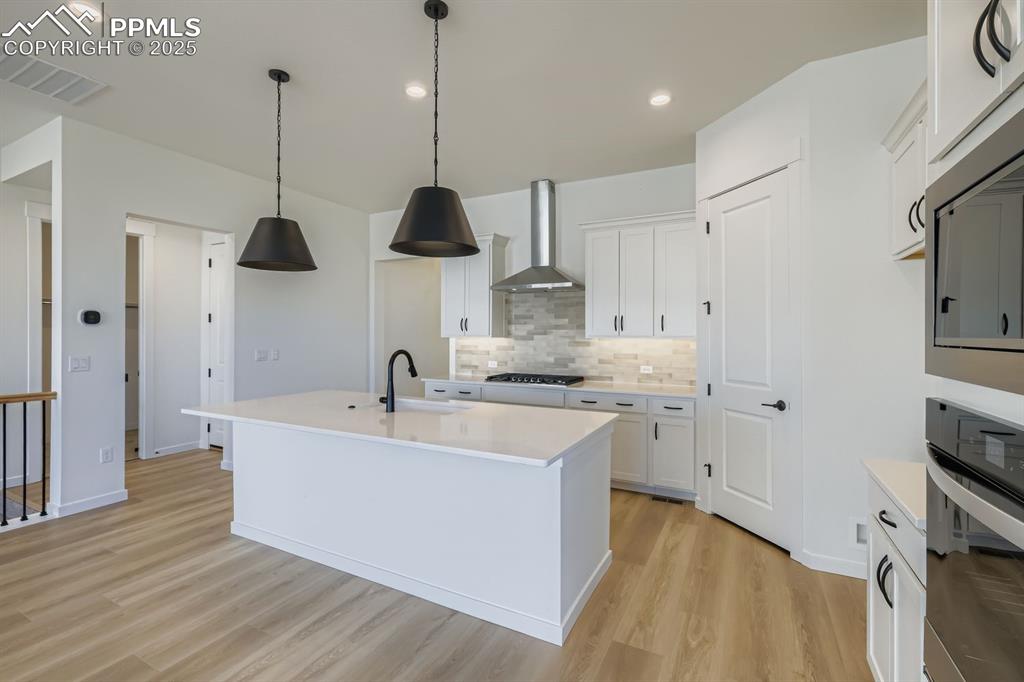 Kitchen featuring a kitchen island with sink, decorative backsplash, stainless steel appliances, light wood finished floors, and white cabinets