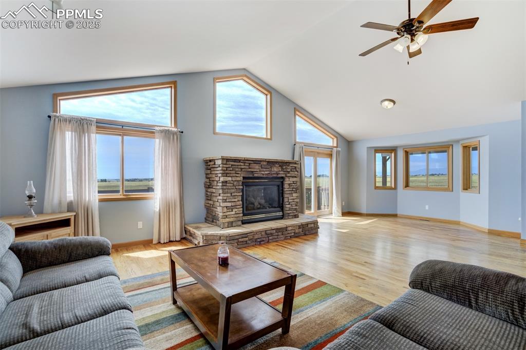 Living area with a stone fireplace, healthy amount of natural light, light wood-type flooring, a ceiling fan, and high vaulted ceiling