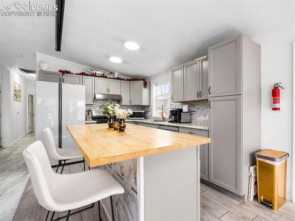 Kitchen with butcher block countertops, gray cabinetry, decorative backsplash, freestanding refrigerator, and a kitchen bar