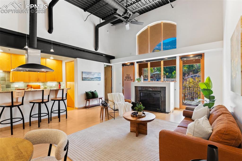 The living area featuring light wood-style flooring, ceiling fan, a tiled fireplace, and an industrial accent ceiling