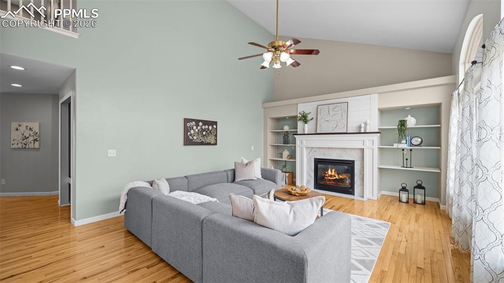 Living room with a high ceiling, a glass covered fireplace, a ceiling fan, light wood finished floors, and built in shelves