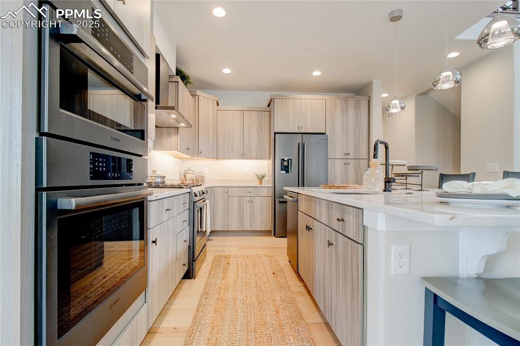 Kitchen featuring light brown cabinetry, appliances with stainless steel finishes, wall chimney range hood, recessed lighting, and an island with sink