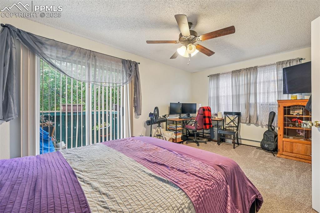 Carpeted bedroom with a textured ceiling, access to exterior, a desk, a ceiling fan, and a baseboard heating unit
