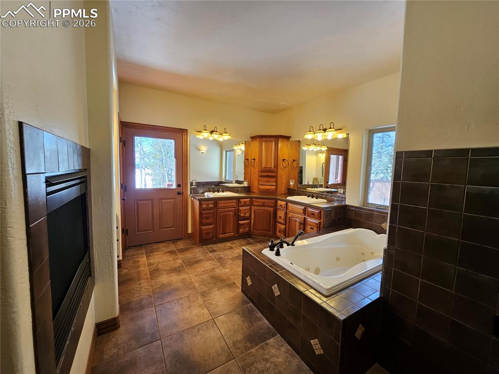 Primary bathroom with two vanities, a jetted tub, healthy amount of natural light, and see-through fireplace