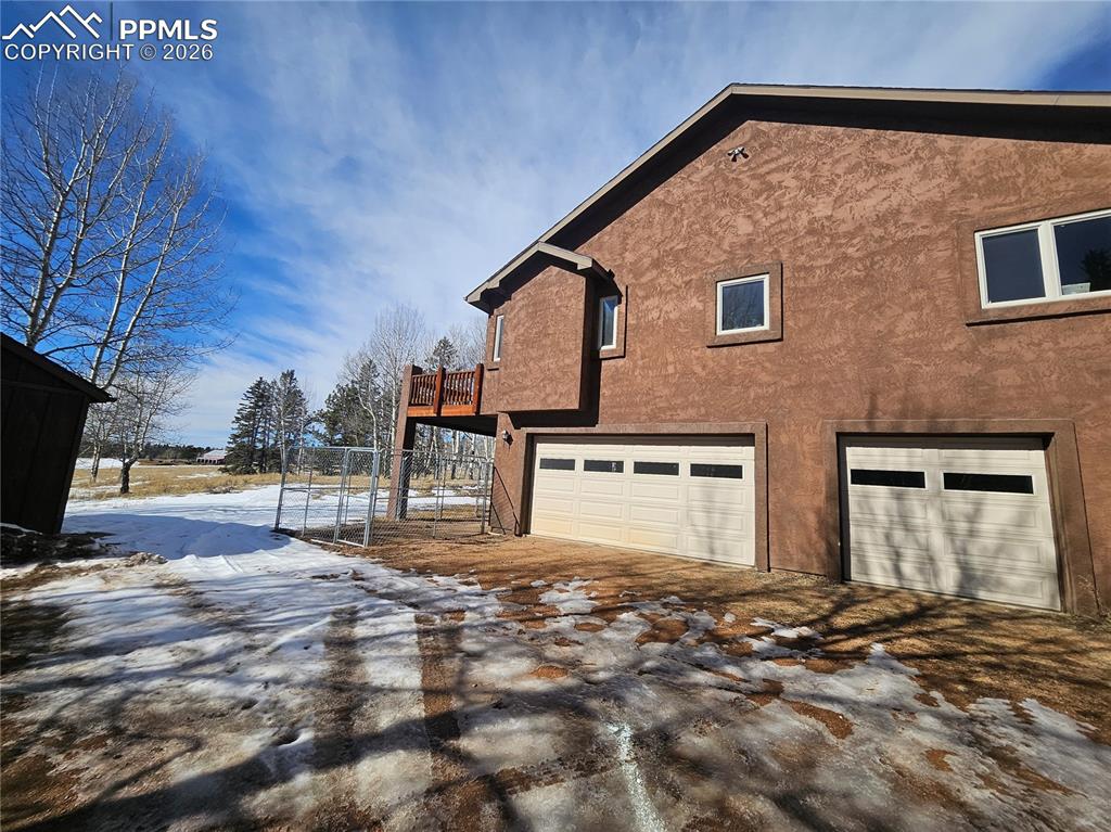 Snow covered gravel driveway with stucco siding, a garage, and a deck - additional large shed to left