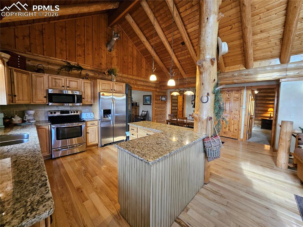 Cabin kitchen featuring log walls, stainless steel appliances, a high wooden beamed ceiling, light stone counters, and decorative light fixtures