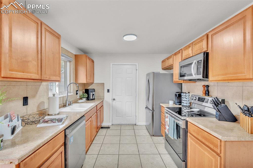 Kitchen featuring stainless steel appliances, backsplash, light countertops, light tile patterned floors, and light wood finish cabinetry