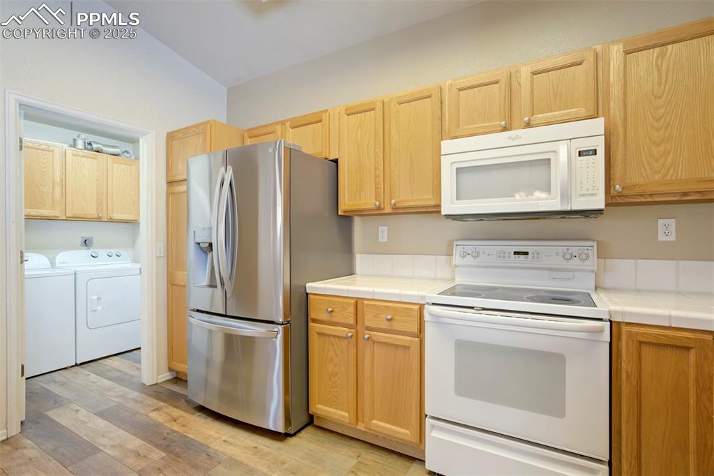 Kitchen with white appliances, light wood finished floors, washing machine and dryer, tile countertops, and light brown cabinets