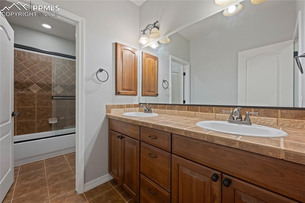 Basement Bathroom with bath / shower combo with glass door, dark tile patterned flooring, and double vanity
