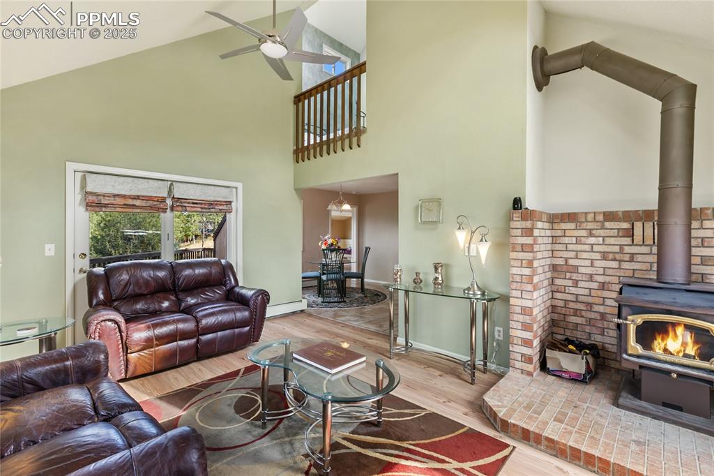 Living room featuring wood finished floors, a wood stove, high vaulted ceiling, ceiling fan, and a baseboard heating unit