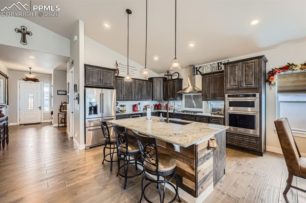 Kitchen with stainless steel appliances, wall chimney exhaust hood, a sink, dark brown cabinetry, and lofted ceiling