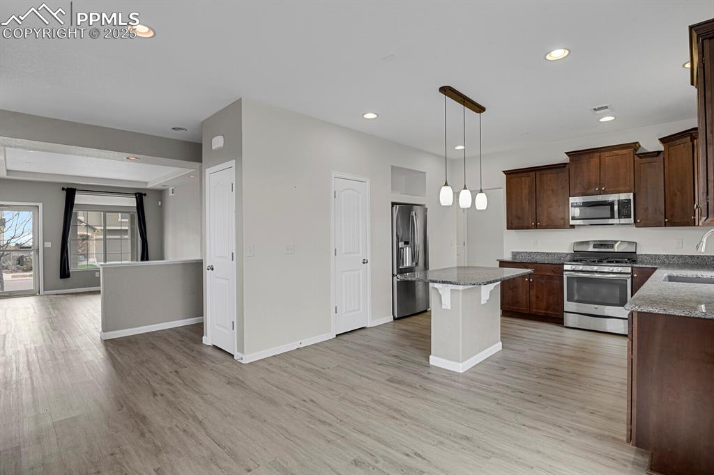 Kitchen with appliances with stainless steel finishes, hanging light fixtures, dark brown cabinetry, dark stone counters, and open floor plan