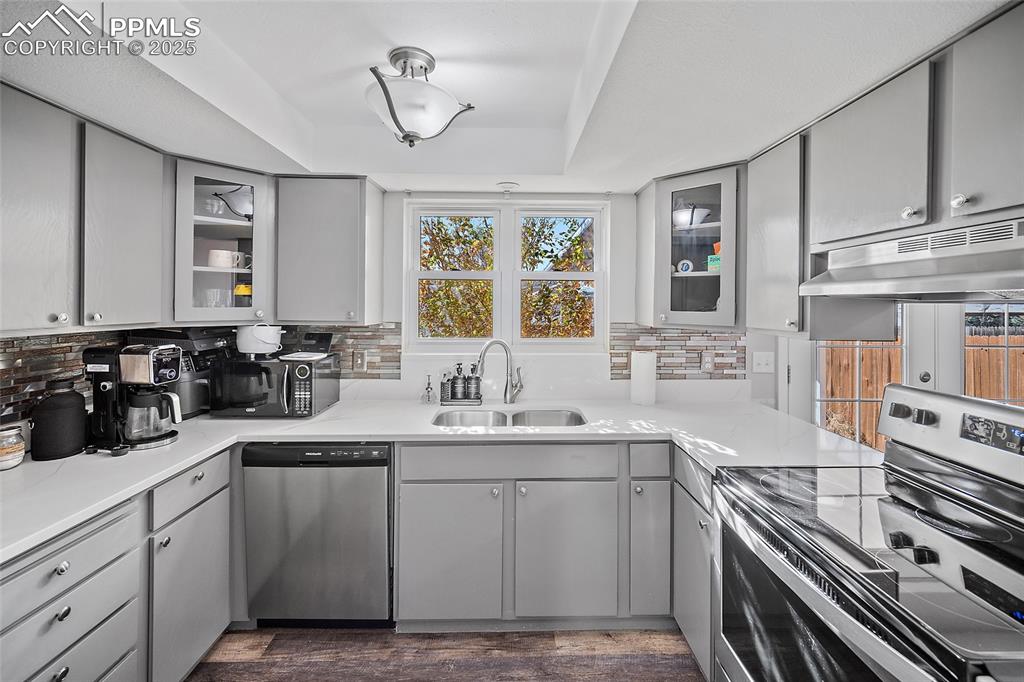 Kitchen featuring gray cabinetry, glass insert cabinets, appliances with stainless steel finishes, under cabinet range hood, and a tray ceiling