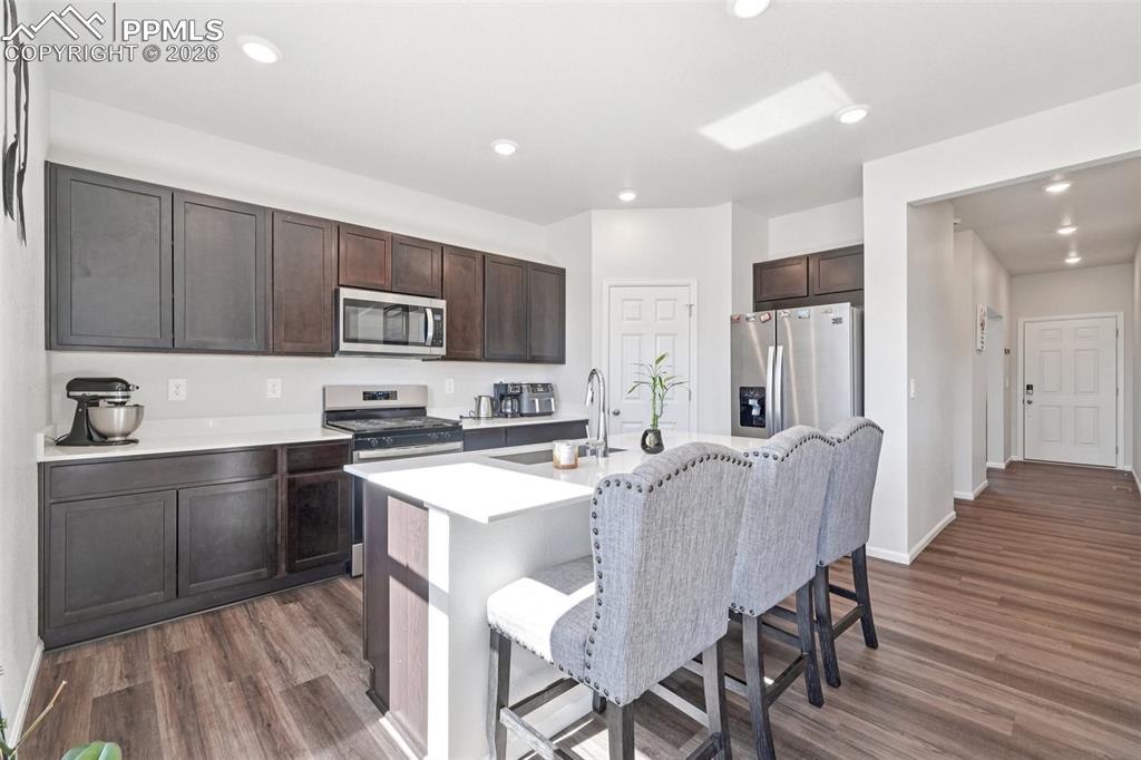 Kitchen with stainless steel appliances, an island with sink, dark wood-style flooring, dark wood finish cabinets, and a kitchen bar