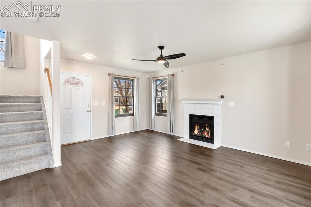 Unfurnished living room featuring wood finished floors, ceiling fan, a tiled fireplace, and stairs