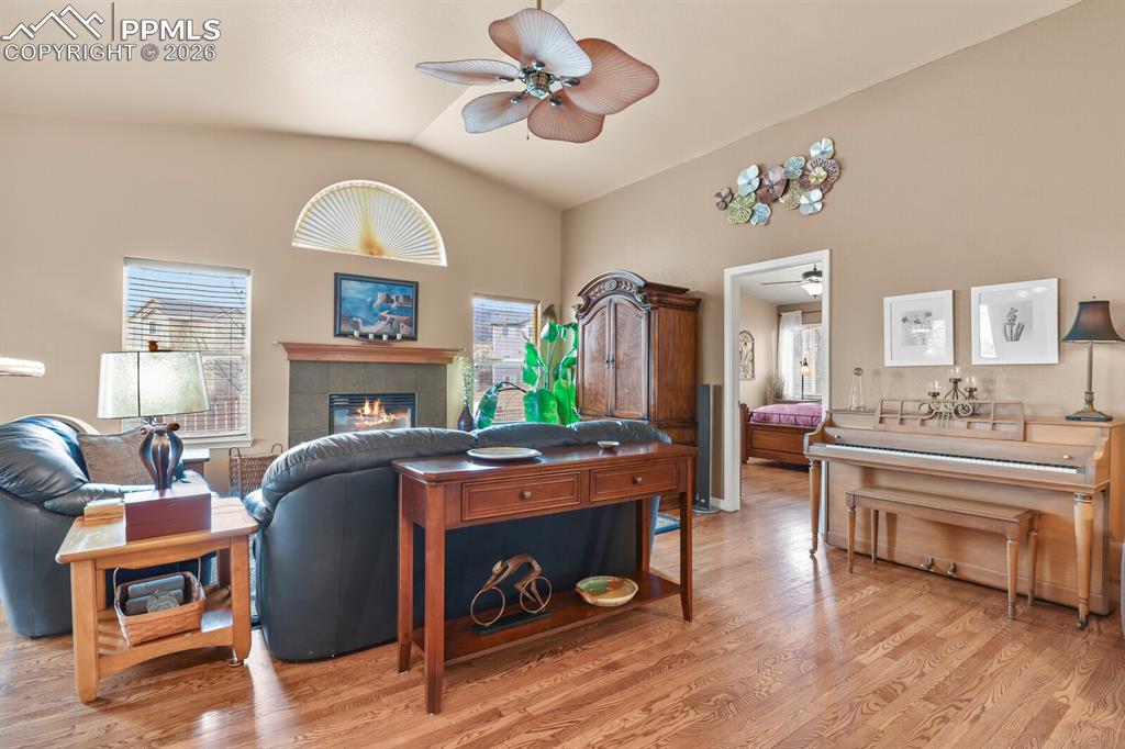 Living room with ceiling fan, wood flooring, vaulted ceiling, a tiled surround to the gas fireplace, and plenty of natural light. Door into the primary bedroom suite.