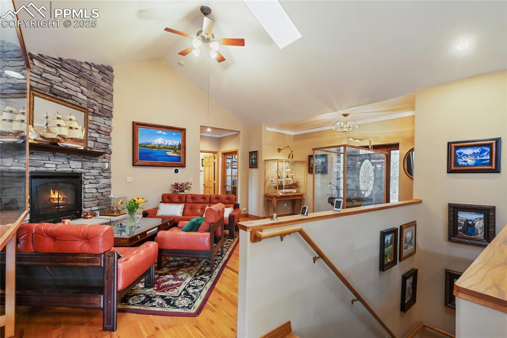 Living room featuring wood-finished floors, a skylight, high vaulted ceilings, a stone fireplace, and a ceiling fan.